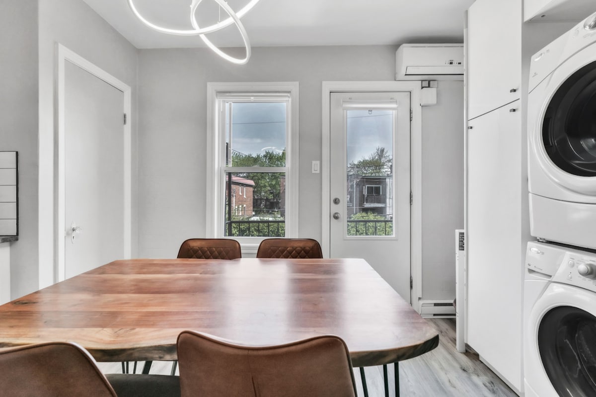A dining area features a wooden table surrounded by four chairs. A large window allows natural light to enter, providing a view of the outdoors. A door leads to a small balcony, while a washer and dryer are located nearby in sleek, white cabinetry.