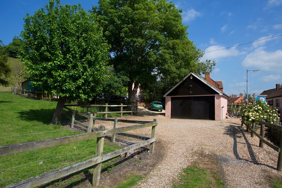 A gravel driveway leads to a pink detached house, framed by lush green trees and a serene landscape. A fenced area with greenery surrounds the property, providing a peaceful atmosphere. The clear blue sky complements the inviting exterior of the home.