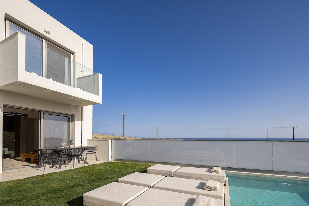 A modern villa exterior is showcased, featuring a sparkling pool surrounded by cushioned sun loungers. The spacious terrace offers access to an open-plan living area, complemented by clear views of the serene sea and sky. Soft green grass extends around the pool area.