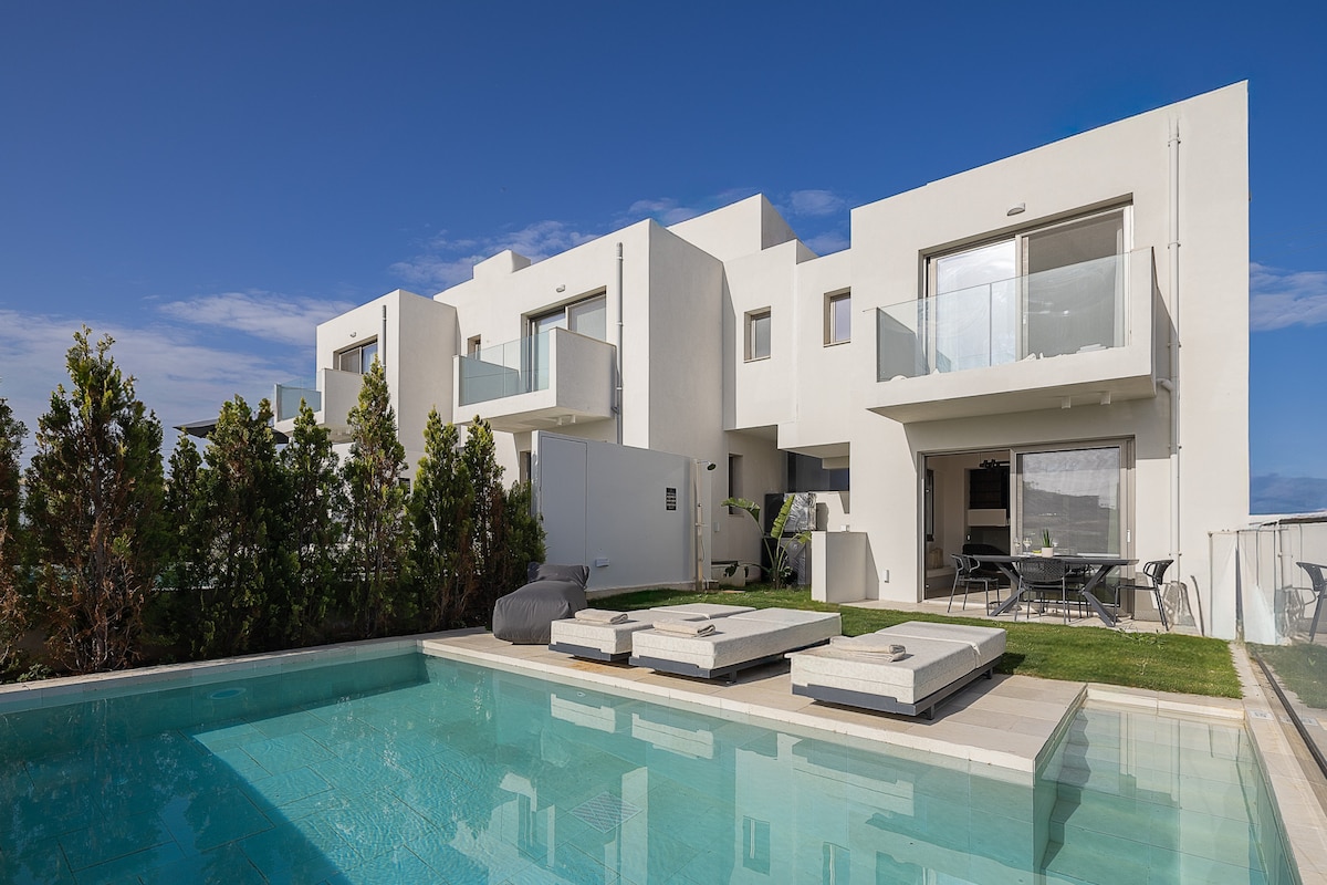 The villa's exterior showcases a private pool surrounded by modern loungers and a well-kept garden. The white-walled structure presents multiple balconies and open living spaces, reflecting a contemporary design against a clear blue sky.