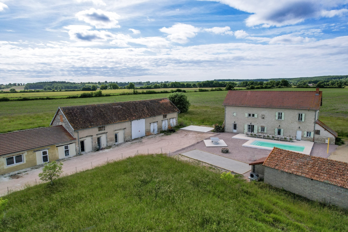 A spacious villa is surrounded by expansive green fields under a partly cloudy blue sky. The modern structure features a poolside area with a rectangular swimming pool and a large patio space, creating an inviting environment for outdoor activities.