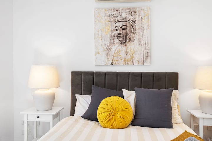 Ugodna spavaća soba s udobnim krevetom, noćnim svjetiljkama i elegantnim umjetničkim djelom koje prikazuje Buddhu. Savršeno za opuštajući boravak.