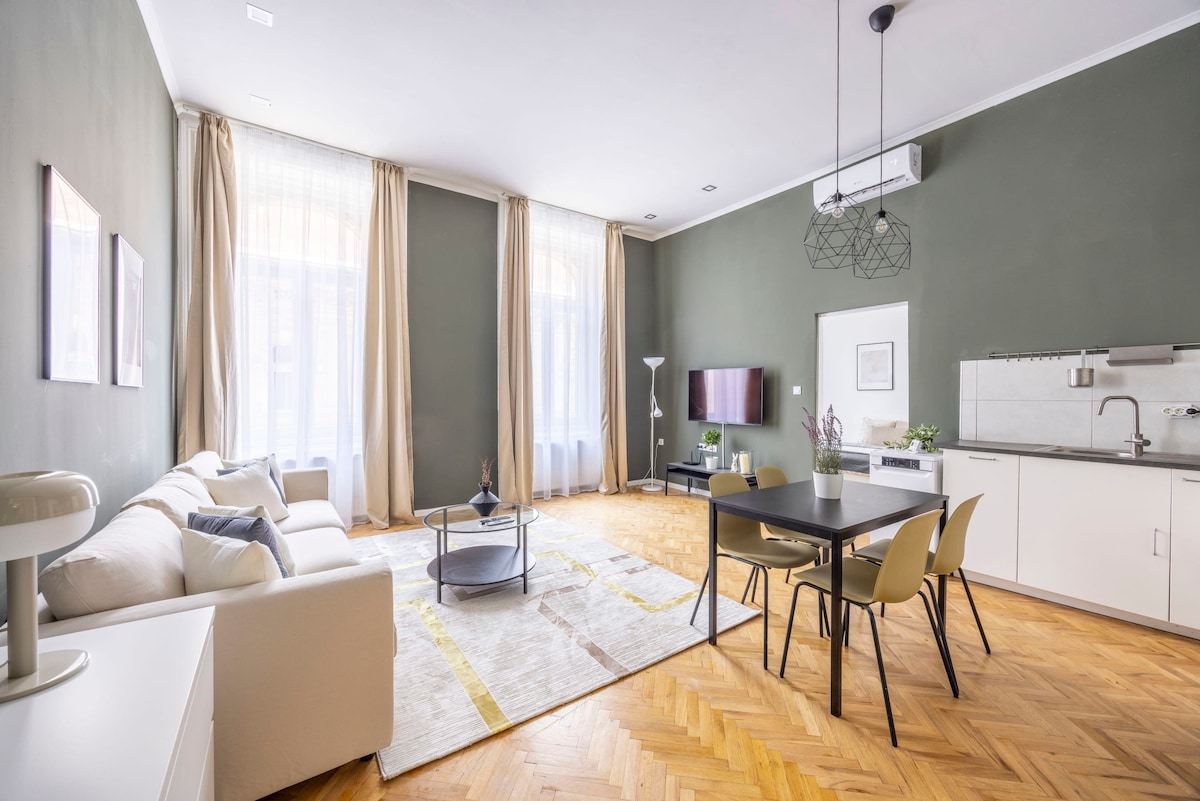 A light-filled living area features high ceilings and large windows, enhancing the sense of space. A comfortable sofa and a round coffee table are positioned on a patterned rug. A dining table with chairs is nearby, and a flat-screen TV is mounted on the wall.