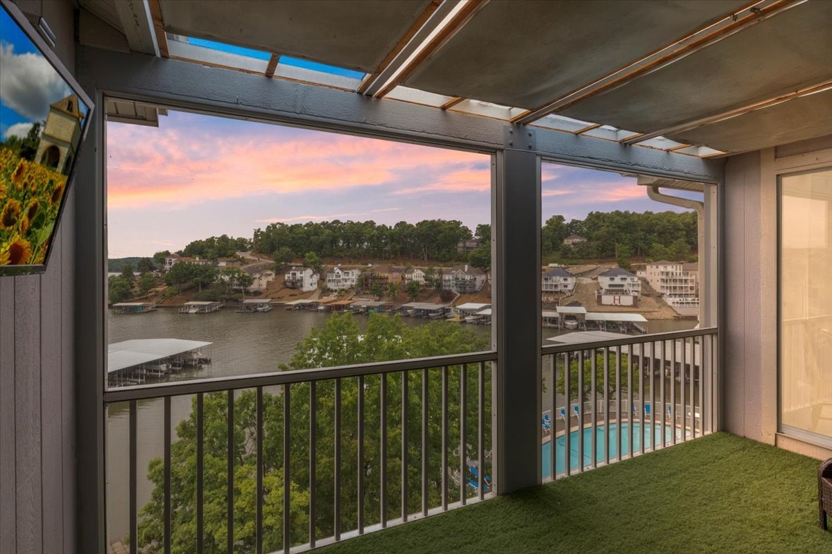 The Astroturf patio features a scenic view of the lake and community pool, framed by expansive windows. A large TV is mounted on the wall, providing entertainment options. The horizon showcases a soft sunset gradient, enhancing the tranquil atmosphere of the outdoor space.