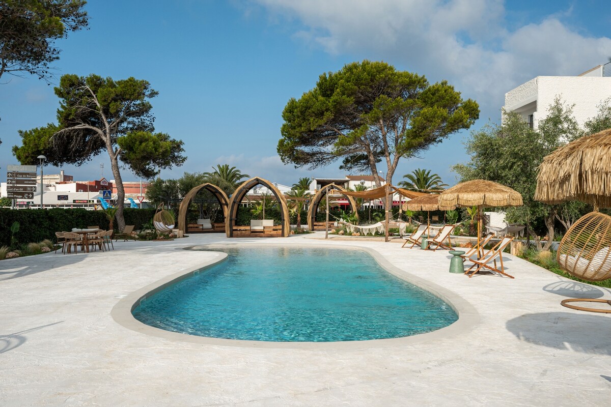 A clear blue pool is surrounded by a spacious terrace featuring stylish lounge chairs and shaded seating areas. The natural landscape includes lush greenery and trees, enhancing the serene environment. Sunlight reflects off the pool surface, creating a peaceful atmosphere for relaxation.