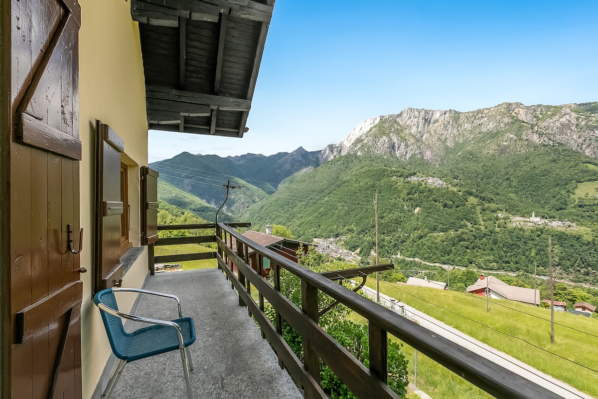 An outdoor balcony features wooden railings, offering a view of the surrounding green mountains and valleys. A single blue chair is positioned on the balcony, providing a space to enjoy the scenic landscape beneath a clear blue sky.