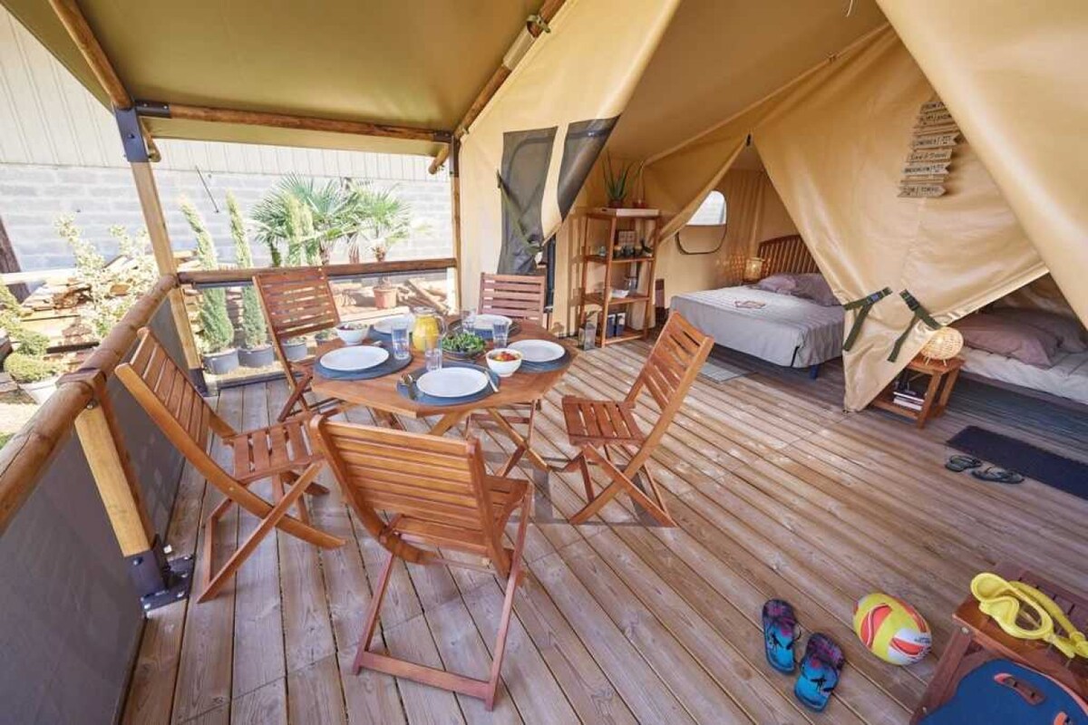 A wooden deck is featured beneath a spacious tent, with a dining table set for four and surrounded by matching folding chairs. A bed is positioned in the background along with a small bookshelf and decorative elements. Bright outdoor shoes are visible on the floor nearby.