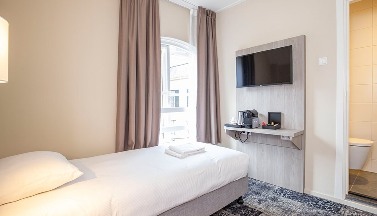 A cozy room features a single bed topped with crisp linens and neatly arranged towels. A wall-mounted TV is positioned above a small table with a kettle. Natural light enters through a window covered by soft curtains, enhancing a calming atmosphere.