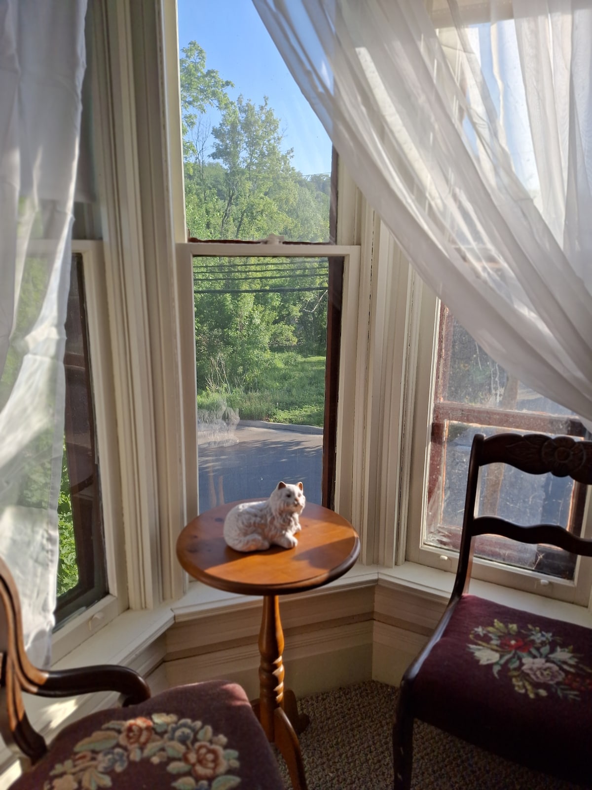 A cozy sitting area is featured in the tower with large windows providing views of the lush greenery outside. A small table holds a decorative figurine, while two chairs with floral upholstery accompany the table, creating an inviting corner for relaxation.