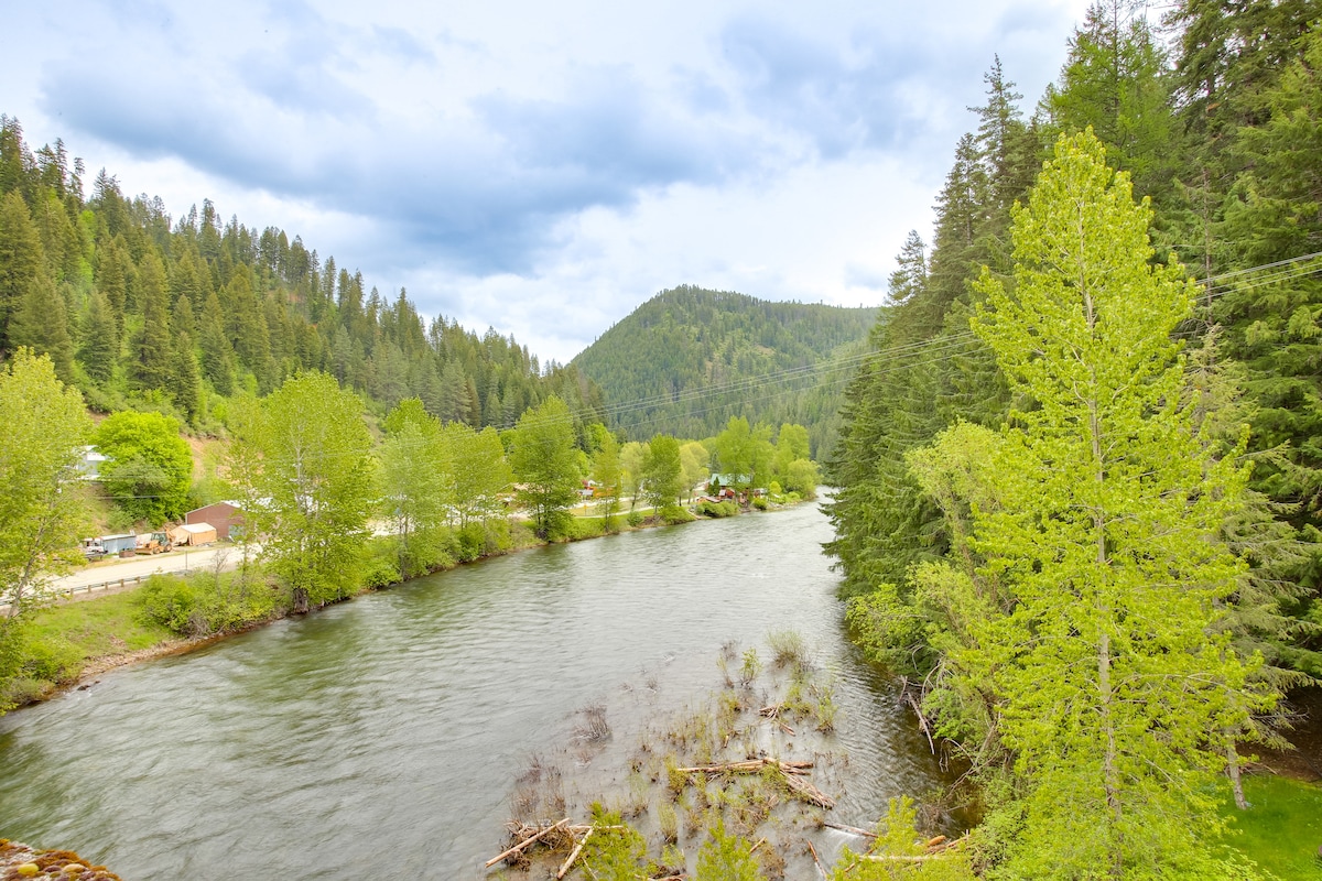 The image captures the serene Saint Joe River, flanked by lush greenery and trees. Rolling hills rise in the background, contrasting with the gentle current of the river. A mix of overcast and clear skies adds to the tranquil atmosphere of this natural setting.