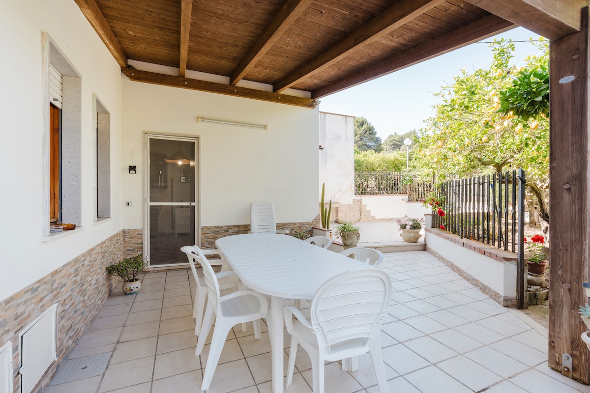 A spacious outdoor terrace is covered by a wooden roof, featuring a large white table surrounded by six white chairs. Potted plants are arranged along the walls, and a garden can be seen in the background, providing a serene environment for relaxation.