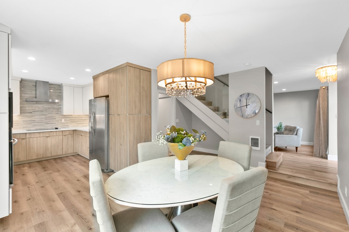 A bright dining area features a circular marble table surrounded by four soft, upholstered chairs. A stylish chandelier hangs above, providing illumination. In the background, a modern kitchen with wooden cabinetry and stainless steel appliances is visible, while a staircase leads to the upper levels.