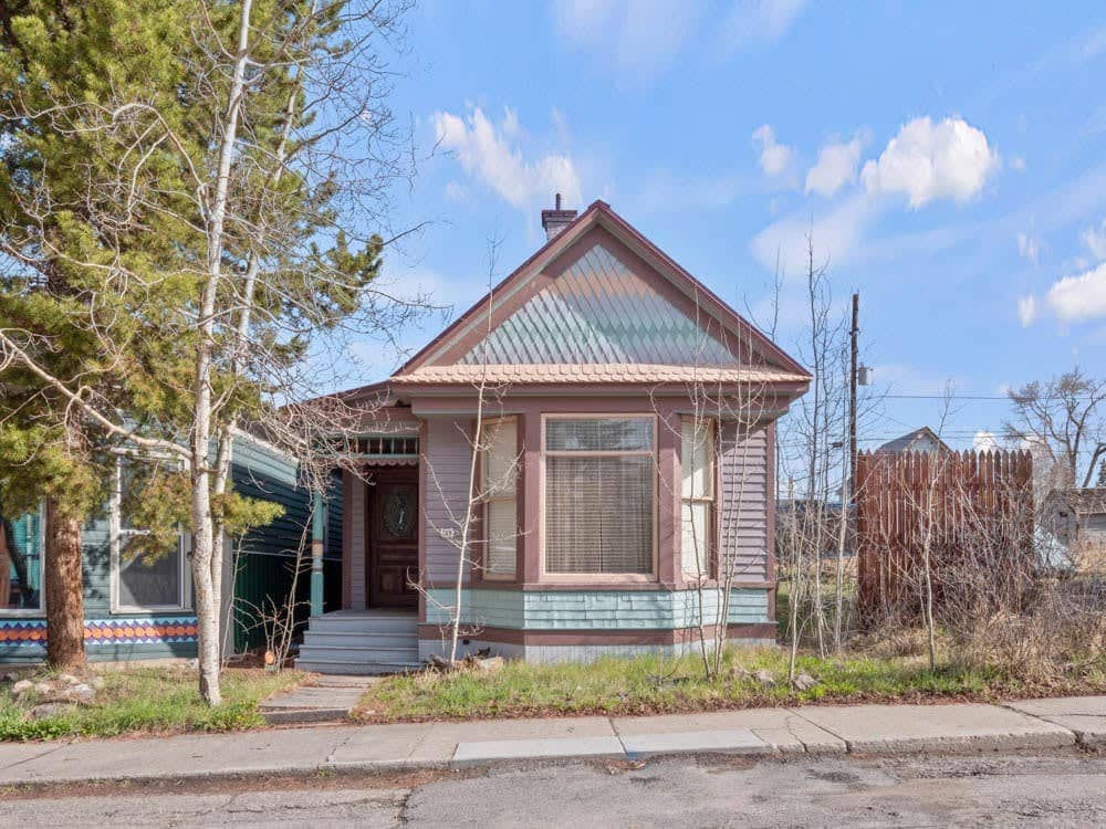 A charming exterior of a historic home is showcased, featuring a peaked roof with multi-colored metal accents. The front porch is adorned with steps leading to a large bay window, framed by shrubs and trees. The surrounding area includes a fenced yard and a sidewalk.