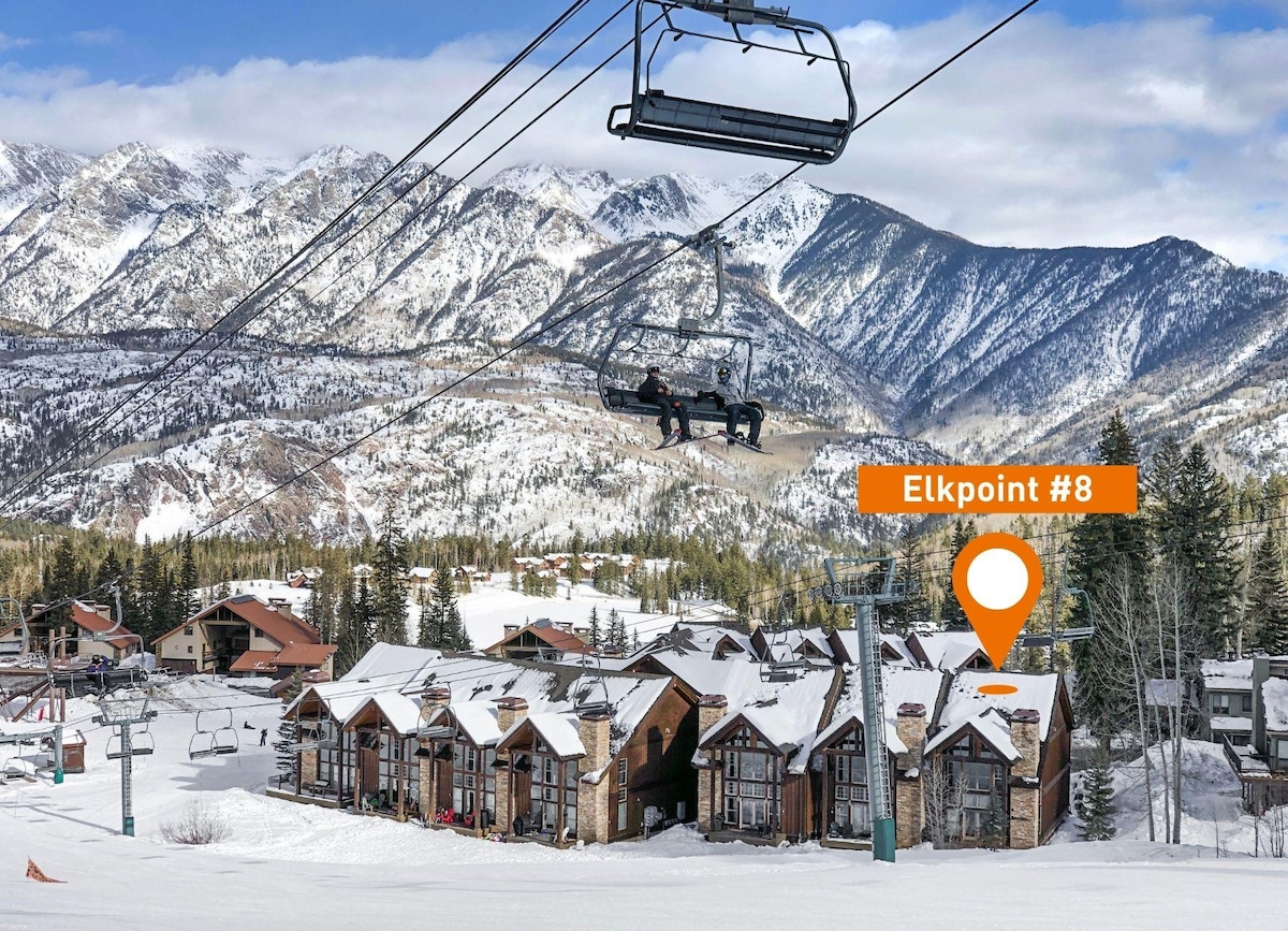 The image displays the exterior of a two-story townhome complex nestled on a snowy slope, with the scenic mountains in the background. A ski lift is seen above, and the location marker points to Elkpoint #8, indicating a ski-in/ski-out property.