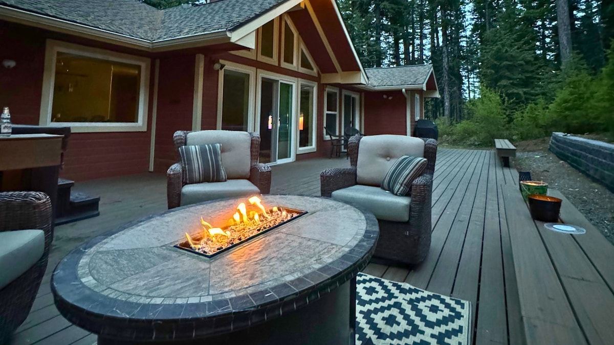 An outdoor deck features a fire pit at the center, surrounded by comfortable seating in woven textures. Large windows provide views into the house, while tall trees create a natural backdrop, enhancing the serene setting. A patterned rug adds subtle detail to the wooden floor.