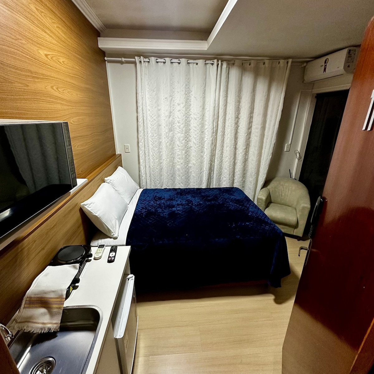 A cozy loft is shown with a spacious bed covered in a blue blanket. A flat-screen TV is mounted on the wall, and a kitchenette is visible with a small refrigerator. A light-colored chair is positioned near a window adorned with white curtains.