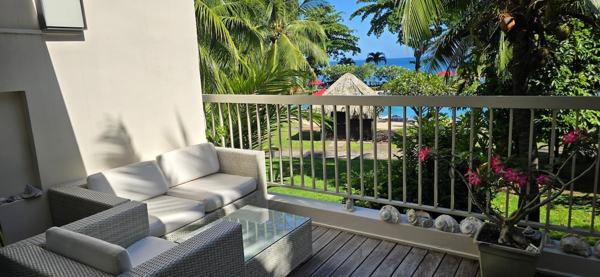 A private terrace features a comfortable seating area overlooking a lush garden and a glimpse of the pool and the sea. Tropical foliage surrounds the space, creating a serene setting for relaxation.