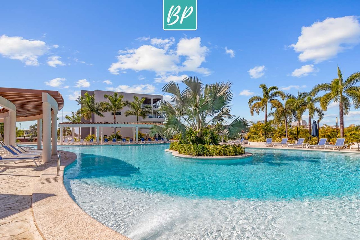 A resort-style pool is surrounded by lush palm trees and lounge chairs, offering a tranquil space for relaxation. The inviting water features a gentle curve, creating a picturesque setting for guests to enjoy. Bright blue skies complement the serene atmosphere.