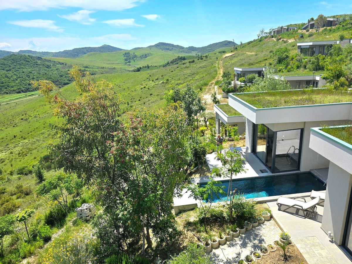 The image captures a modern holiday home nestled in a lush, green landscape. A clear swimming pool reflects the sunlight, surrounded by well-maintained gardens. Scenic hills extend into the horizon, enhancing the natural beauty of the location.