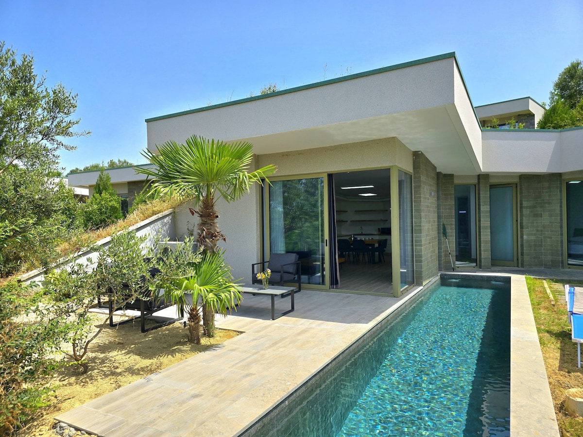 An outdoor space features a private swimming pool with a clear blue surface. Comfortable garden furniture is positioned beside the pool, surrounded by lush greenery and palm trees. The modern holiday home showcases large glass doors that open to the inviting outdoor area.