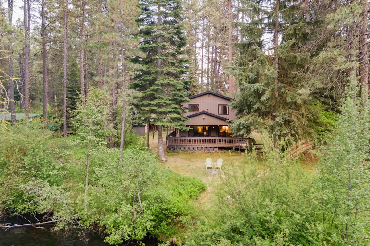 This riverfront home is surrounded by lush greenery and tall trees, offering a serene retreat. The two-story cabin features a spacious deck with seating, seamlessly blending with the natural landscape. The Metolius River is visible nearby, enhancing the tranquil setting.