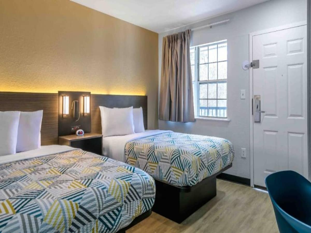A brightly lit room features two full beds with colorful patterned bedding. Each bed is accompanied by modern bedside lamps. A generous window allows natural light to fill the space, while a blue chair and a door leading outside are visible.
