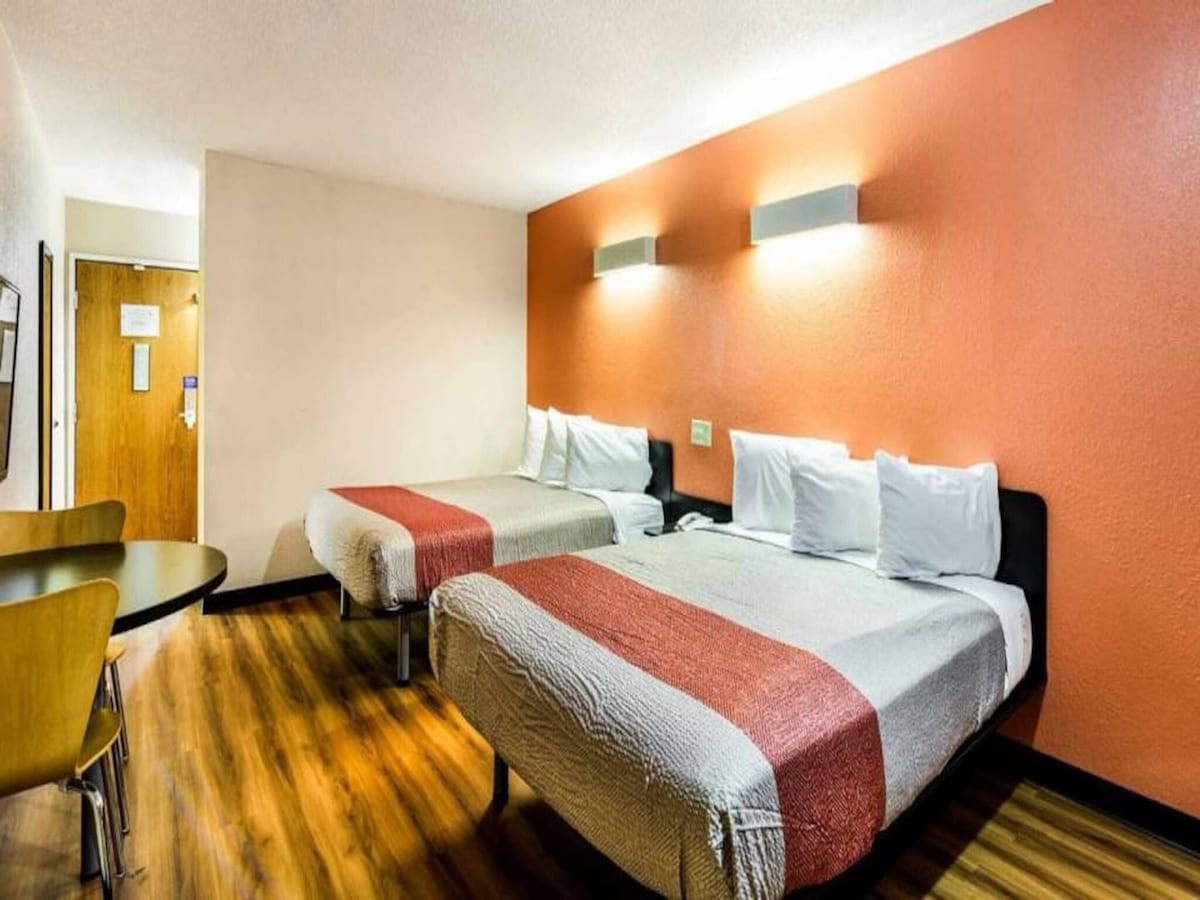A room featuring two queen beds with white bedding and decorative throws. An accent wall in a muted orange tone adds warmth. A small round table with wooden chairs is positioned beside the entrance. Light fixtures are mounted above the beds, illuminating the space.
