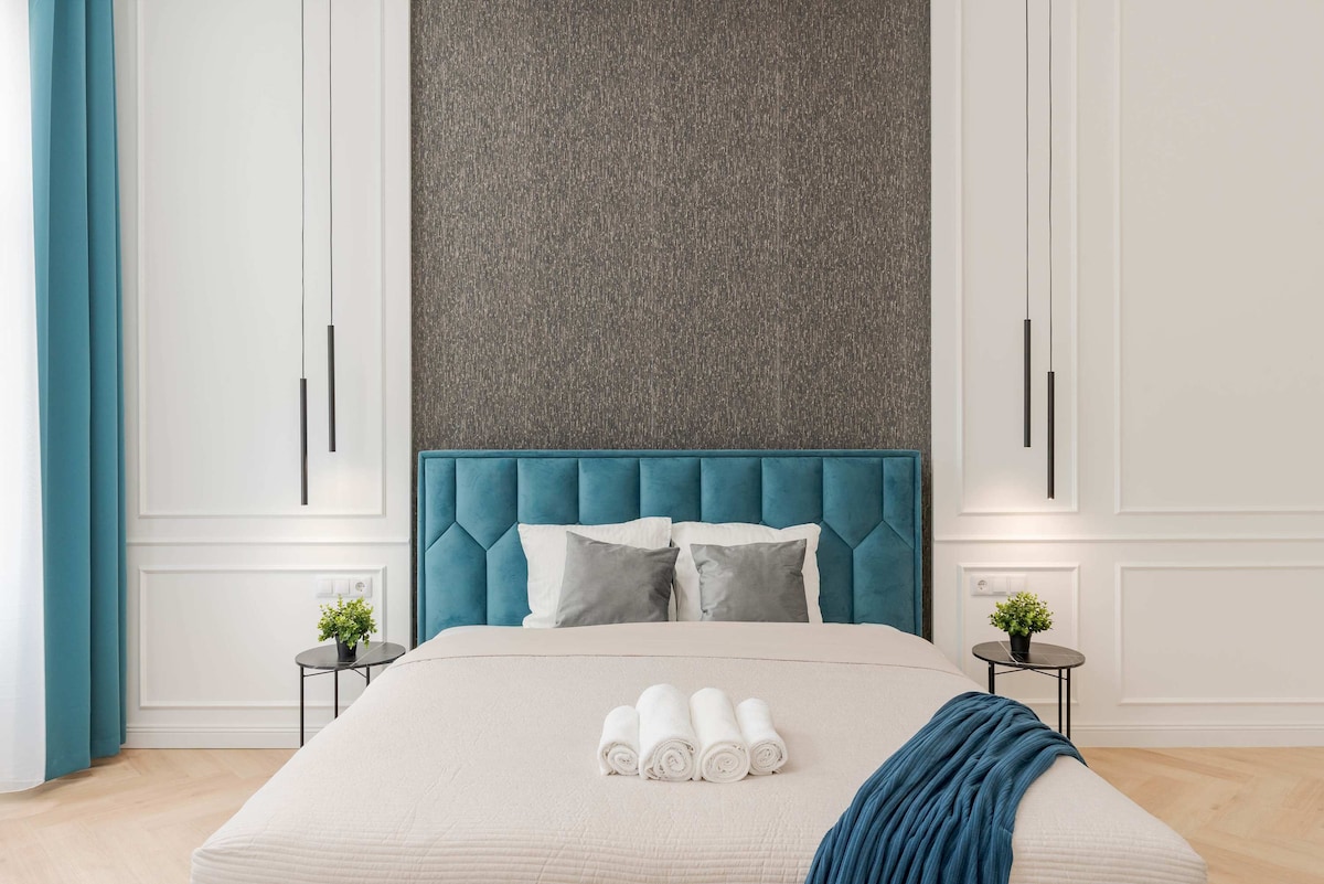 A contemporary bedroom features a plush blue upholstered headboard, complemented by two small side tables with plants. A neutral-colored bedspread covers a spacious bed, adorned with rolled towels. Tall windows are framed by soft blue curtains, allowing natural light to enhance the room.