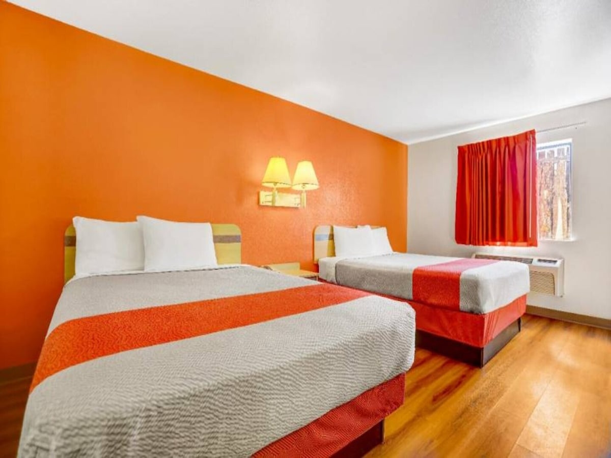 A room features two queen beds dressed in light-colored linens, positioned against an orange accent wall. A window with red curtains allows natural light in, while a nightstand with lamps is located between the beds. Air conditioning and heating units are visible on the wall.