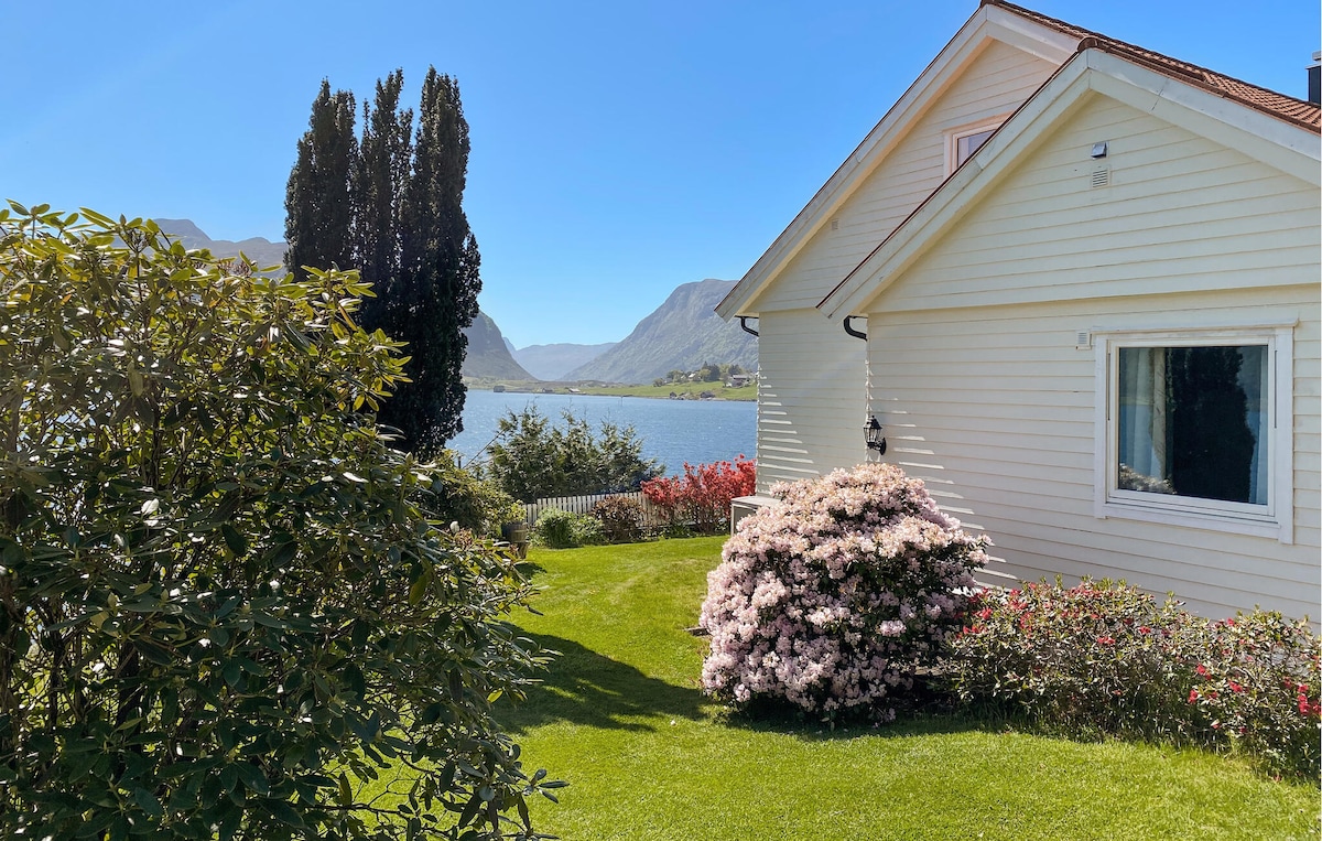 A serene outdoor view features a well-maintained lawn with blooming flowers and shrubs. A white house stands to the right, with a clear window overlooking the water and distant mountains under a bright blue sky.