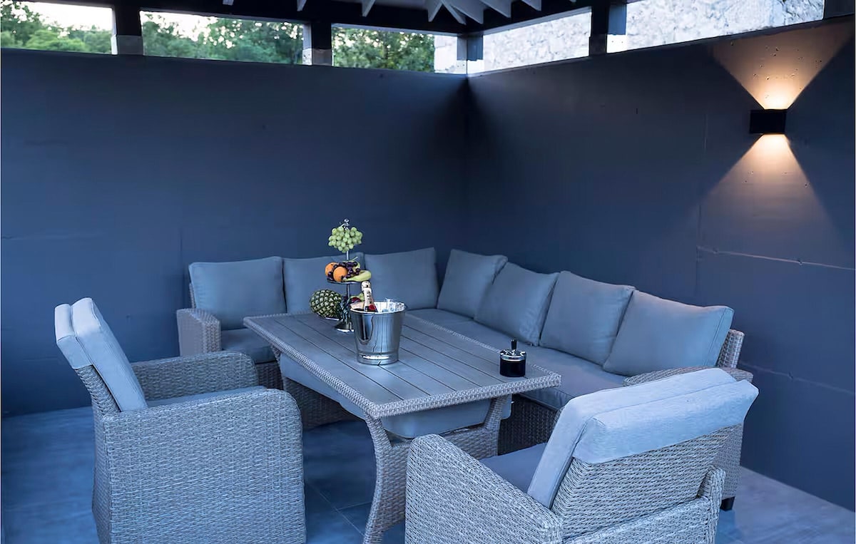A cozy outdoor lounge area is furnished with a large rectangular table surrounded by plush gray seating. Soft lighting illuminates the space, creating a serene environment. The walls feature a smooth, dark finish, enhancing the overall calming atmosphere.