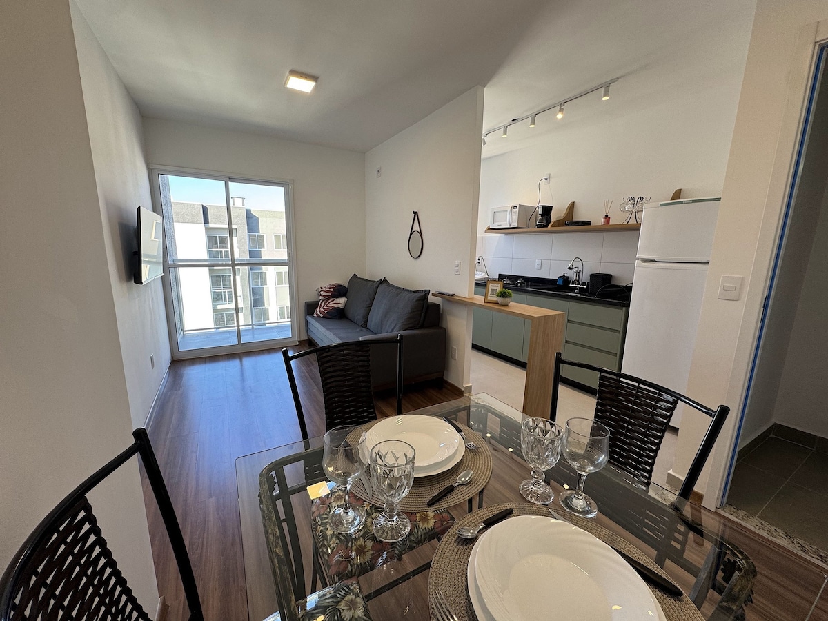 A modern living space features a comfortable sofa adjacent to a dining table set for four. The open kitchen area is visible in the background, showcasing sleek cabinetry and appliances. Large windows allow natural light to fill the room, offering a glimpse of the outdoor view.