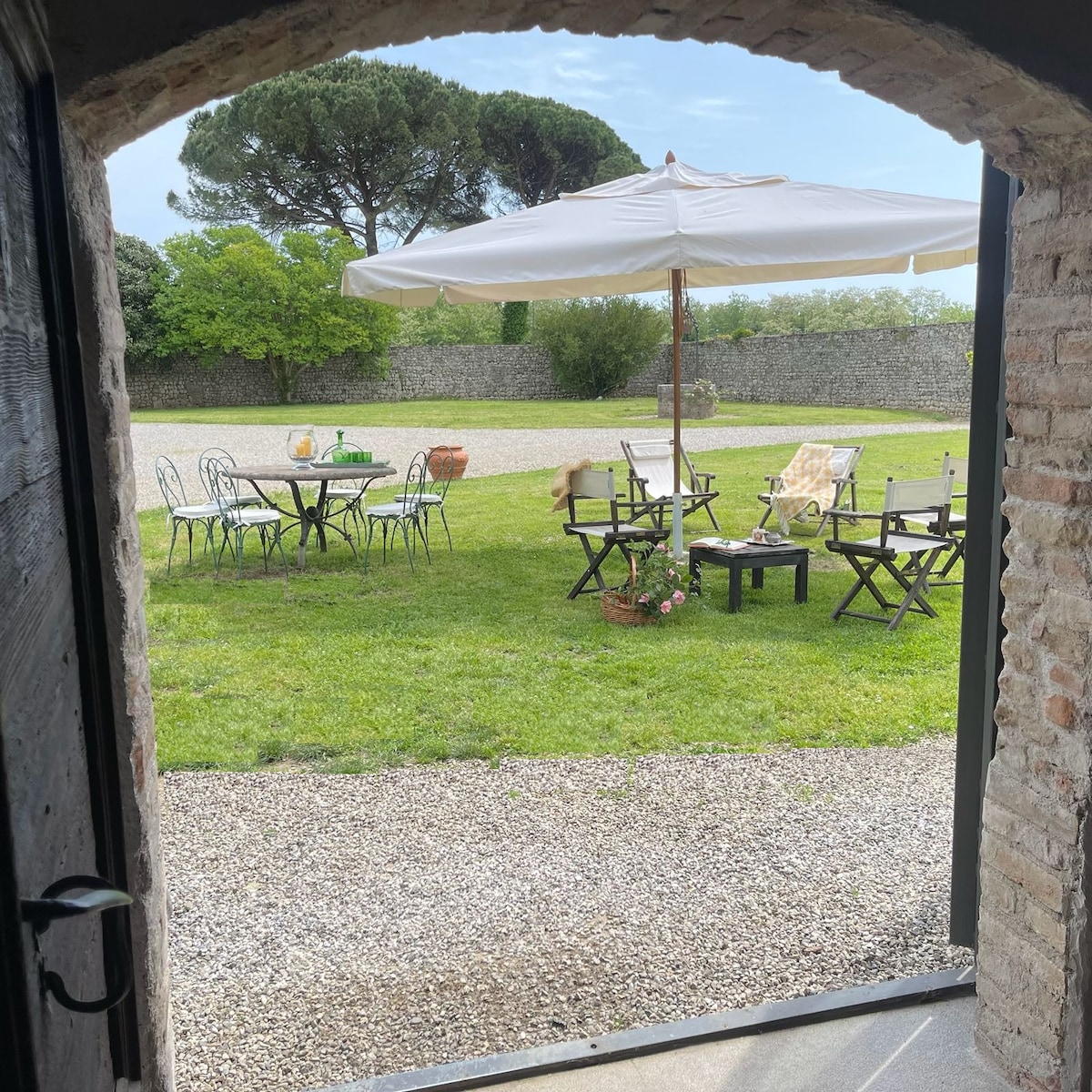 A garden area is visible through a stone archway, featuring a round table with chairs and a large umbrella providing shade. Additional seating, including folding chairs, surrounds the grassy space, which is adorned with potted plants and trees in the background.