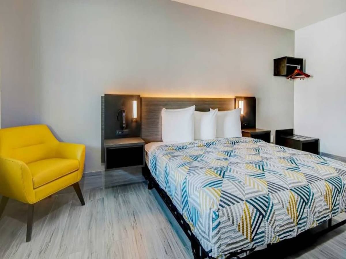 A spacious sleeping area features a king bed with a patterned comforter and two white pillows. A bright yellow chair adds a pop of color beside the bed. Nightstands are positioned on either side, with light fixtures mounted on the wall.