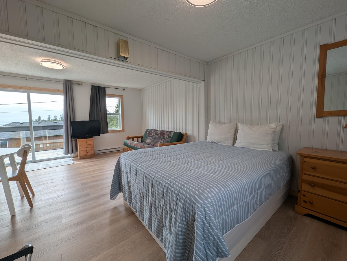A bright and airy suite features a spacious queen bed with a light blue striped blanket. Nearby, a seating area includes a couch and a small table. Natural light fills the room through large sliding doors leading to a deck, offering views of the outdoors.