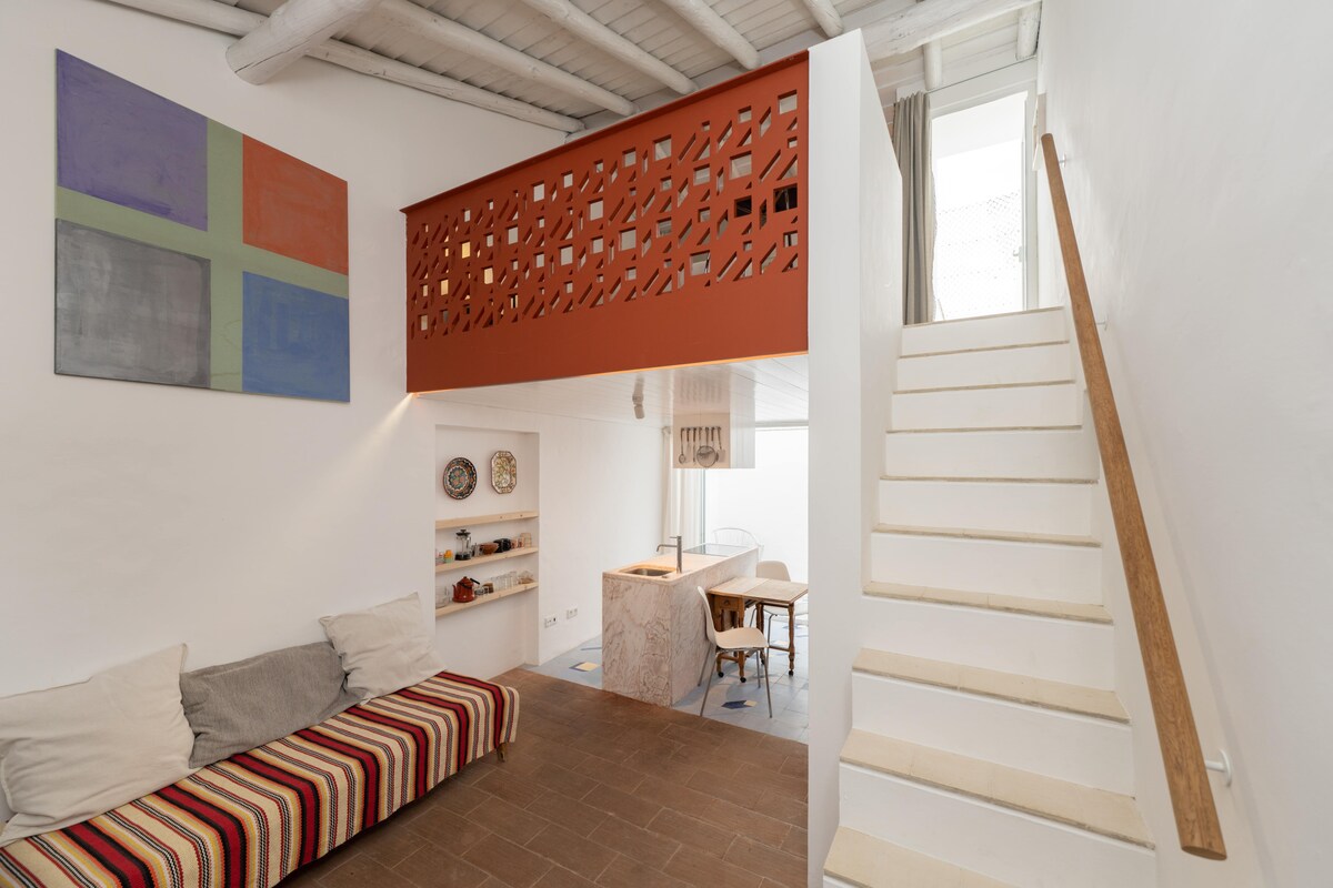 An open living space is visible, featuring a striped sofa accompanied by neutral pillows. A warm orange partition adds a modern touch, while a staircase leads to the upper area. Sunlight filters through large windows, illuminating the minimalist decor and wooden accents.
