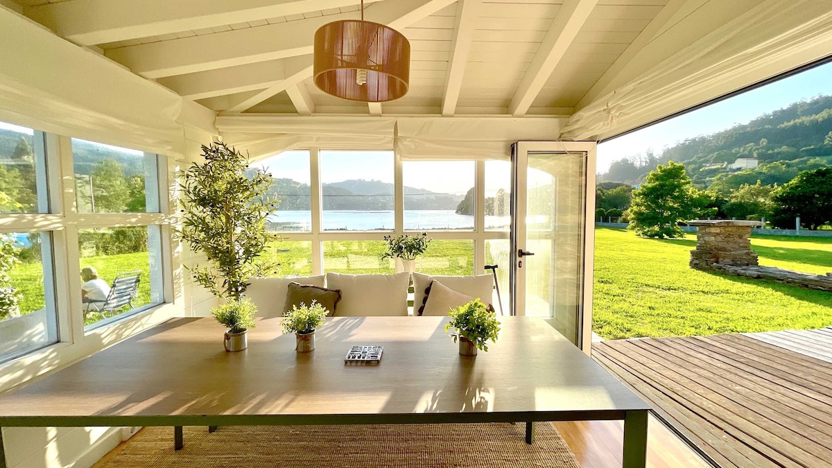 A spacious sunlit gallery features large glass doors opening to a serene view of Cedeira Bay. A brown dining table with plants is centrally placed, complemented by a cozy seating area. Rolling green lawns and distant hills create a natural backdrop visible through the windows.