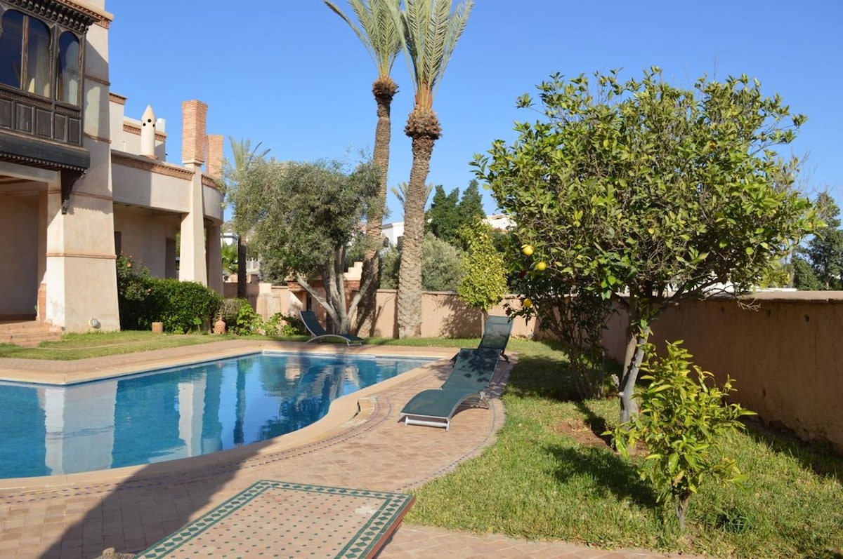 A serene outdoor area features a swimming pool surrounded by a spacious garden. Sun loungers are positioned near the pool, complemented by palm trees and lush greenery. A pathway leads through the garden, enhancing the inviting atmosphere of the space.