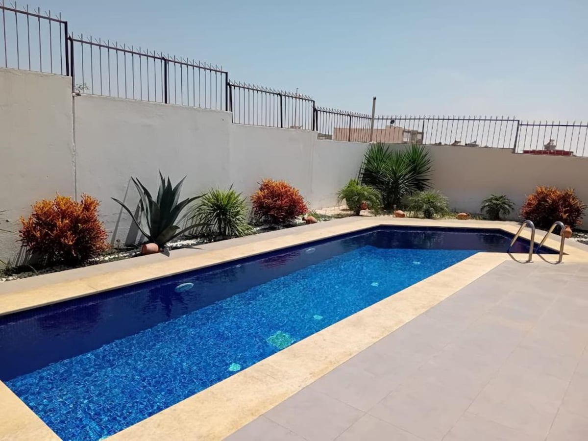 A serene swimming pool is showcased, featuring clear blue water that reflects sunlight. Surrounding plants and flowers are planted along the edges, creating a natural and inviting atmosphere. The pool area is enclosed by a minimalist wall and a metal fence.
