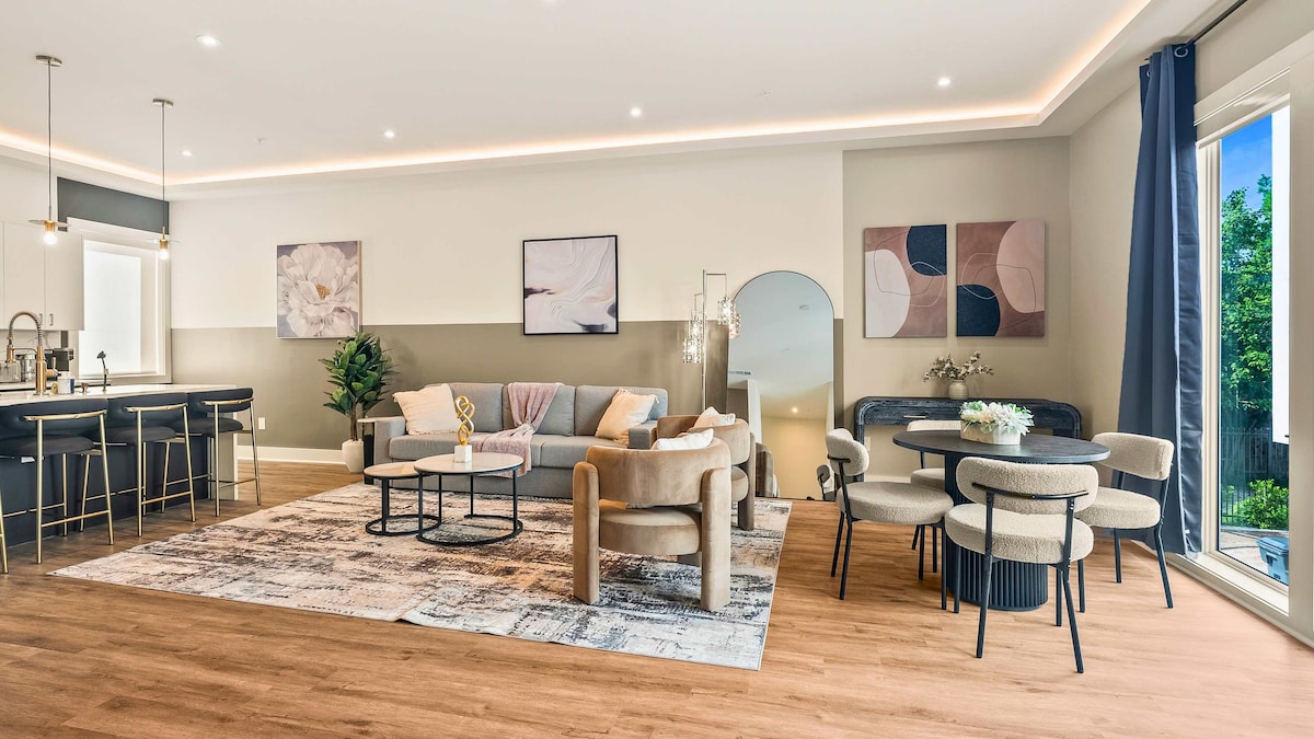 The open-concept living area showcases modern furnishings, including a comfortable sofa and a dining table for six. Bright lighting enhances the space, with contemporary artwork adorning the walls. Large windows invite natural light, while the flooring features warm wood tones.