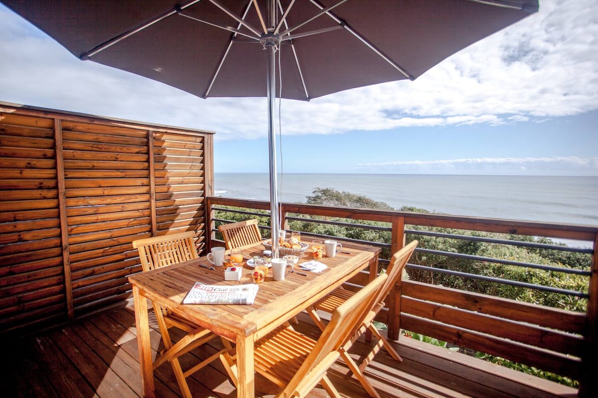 An outdoor dining area features a wooden table surrounded by five chairs, shaded by a large umbrella. The space offers a view of the sea, with lush greenery visible in the foreground and a clear sky above.