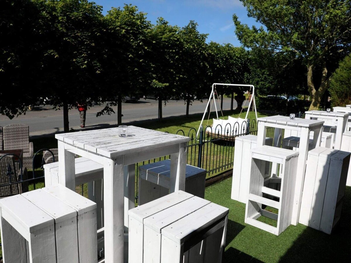 An outdoor area features several white wooden tables and stools arranged on the grass. A swing set is visible in the background, surrounded by leafy trees and a low fence, creating a pleasant setting for relaxation and socializing.