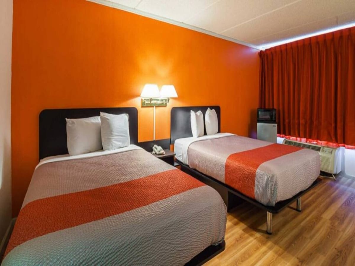 A room is depicted featuring two full beds with grey bedspreads. An orange accent wall complements the vivid red curtains framing a window. Nightstands with lamps are positioned beside the beds, and a small refrigerator is visible in the corner.