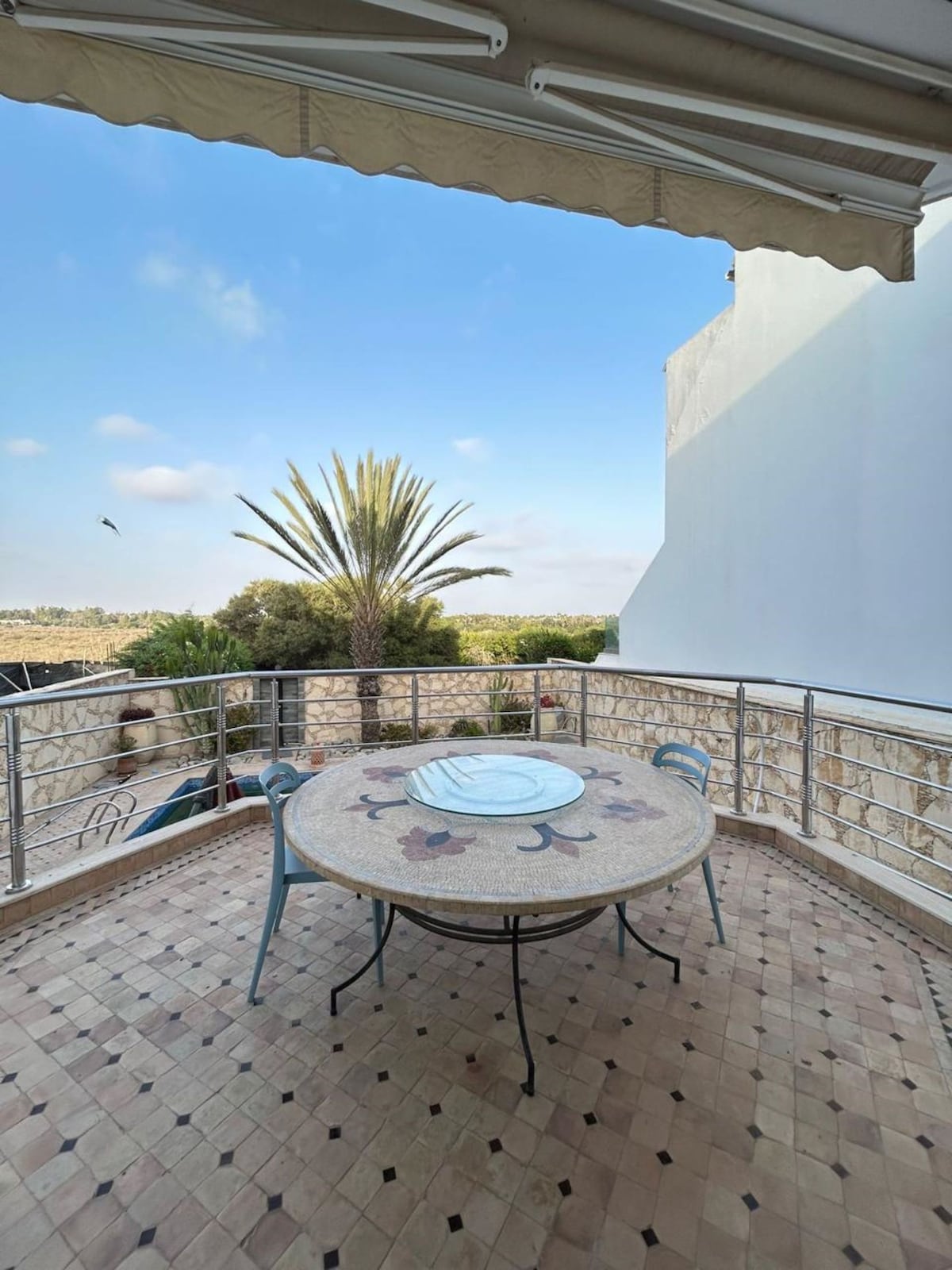 A balcony space features a round table with a decorative mosaic pattern surrounded by four blue chairs. In the background, a tall palm tree stands against a clear sky, with lush greenery visible beyond the railing.