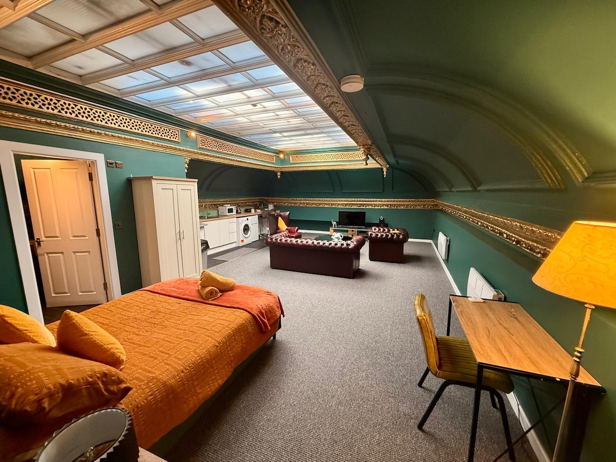 A spacious room is illuminated by natural light from a skylight. It features a double bed adorned with warm orange bedding and cushions, complemented by a cozy sofa area. A small dining table and chair are positioned next to the kitchenette, showcasing modern amenities against a rich green wall.