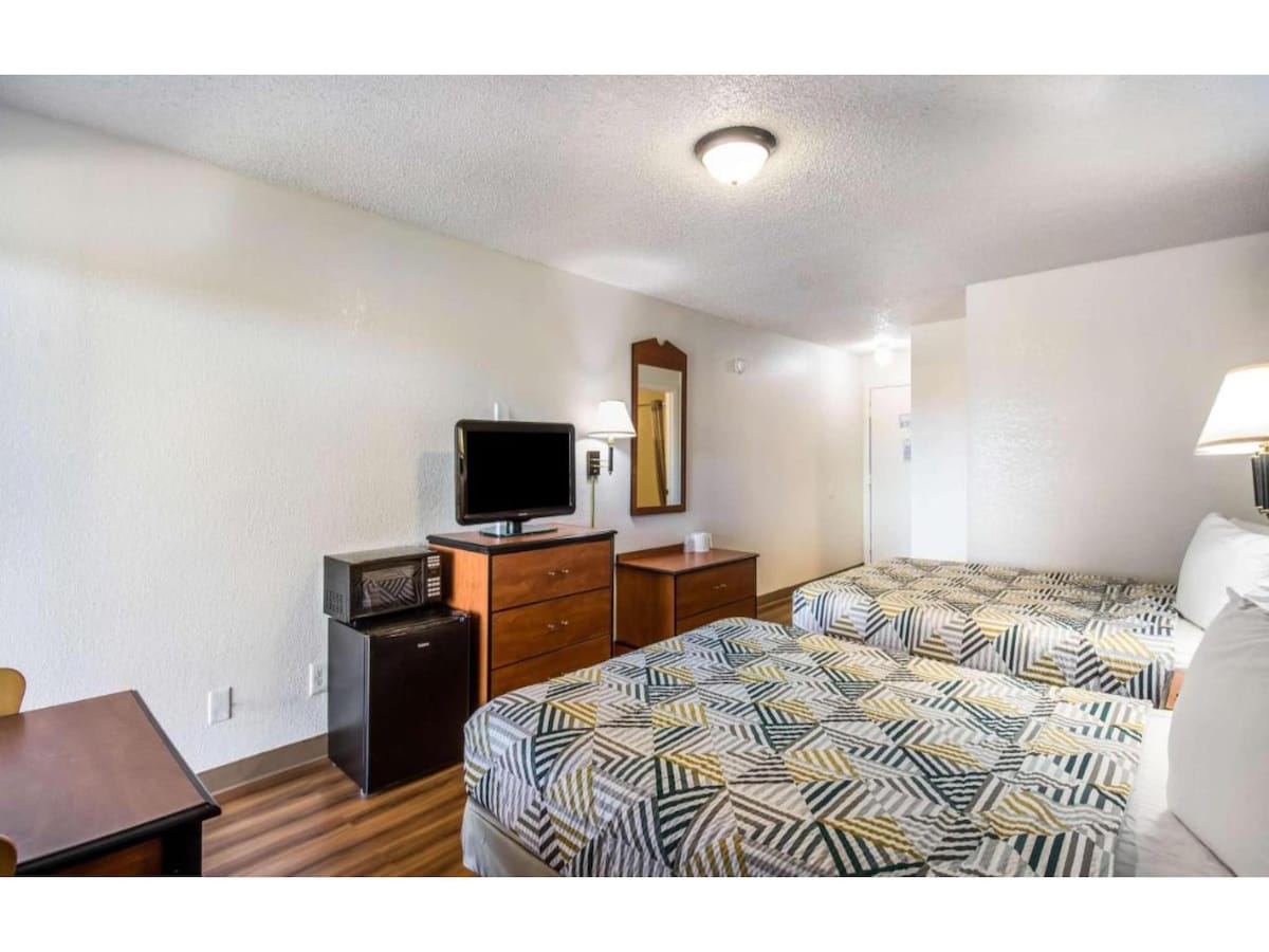 A spacious room features two queen beds dressed in patterned bedding. A television and a microwave sit atop a wooden dresser. A small desk and chair are visible, alongside a light illuminating the serene atmosphere of the area.