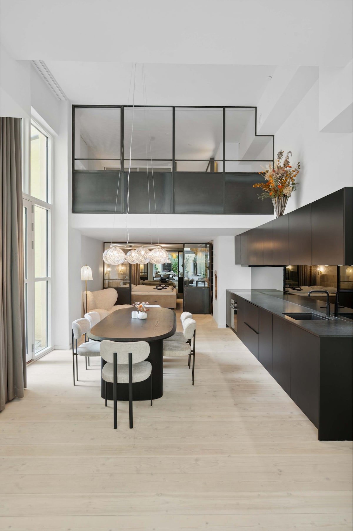 A modern dining area displays a long wooden table surrounded by light-colored chairs. Above, artistic lighting fixtures hang elegantly, while a sleek black kitchen is visible along one wall. Tall windows provide ample natural light, enhancing the spacious and stylish atmosphere.
