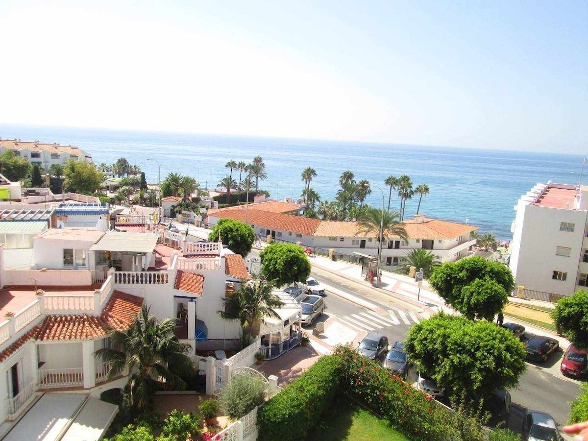 The image captures a vibrant coastal scene overlooking the sea. Lush palm trees and greenery surround the area, with buildings featuring terracotta roofs visible below. Clear blue waters meet the shoreline, extending to the horizon under a bright sky.