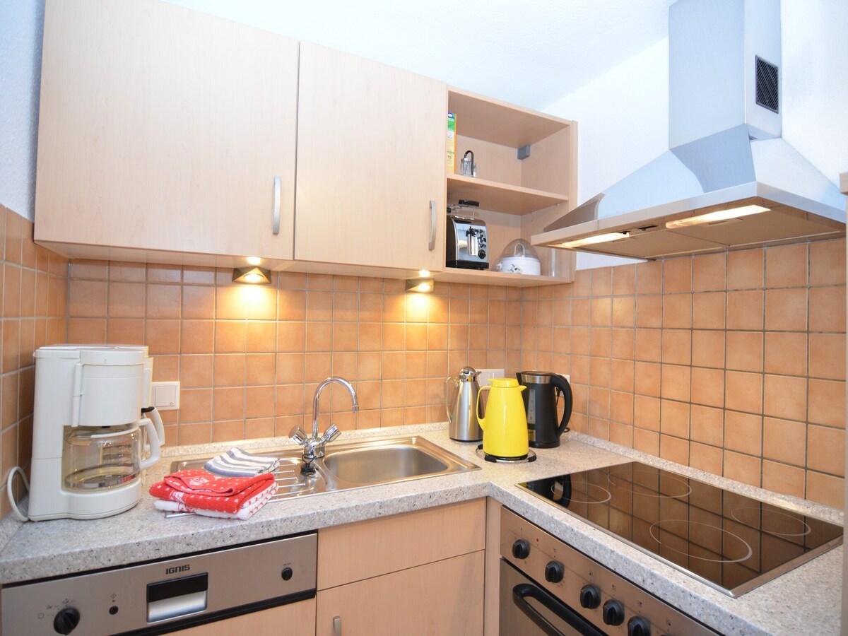The kitchen features a stone countertop with a built-in electric cooktop and oven. A sink is positioned under the cabinetry, complemented by a coffee maker, kettle, and various kitchen utensils on display. Warm lighting enhances the functional space with ample storage.
