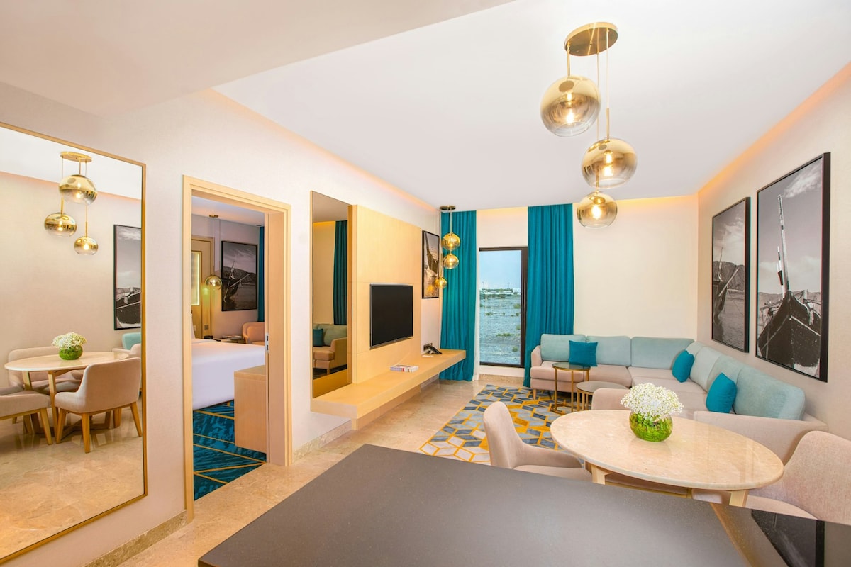 A modern living area features a light-colored seating arrangement, a round dining table, and a wall-mounted television. Decor is complemented by large windows offering views of the outdoors, and golden pendant lights hang from the ceiling, enhancing the stylish ambiance.